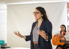 Woman presenting at an indoor business event with a guitar player in the background.