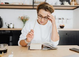 A focused woman in glasses reading a book in a bright, modern kitchen setting with coffee.